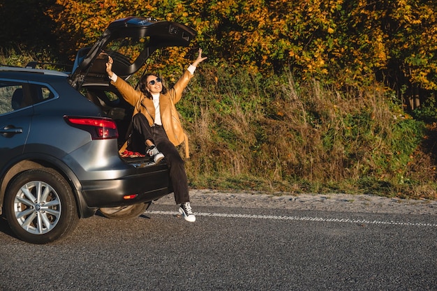 秋、幸せな女性が両手を上げて車のトランクに座っている