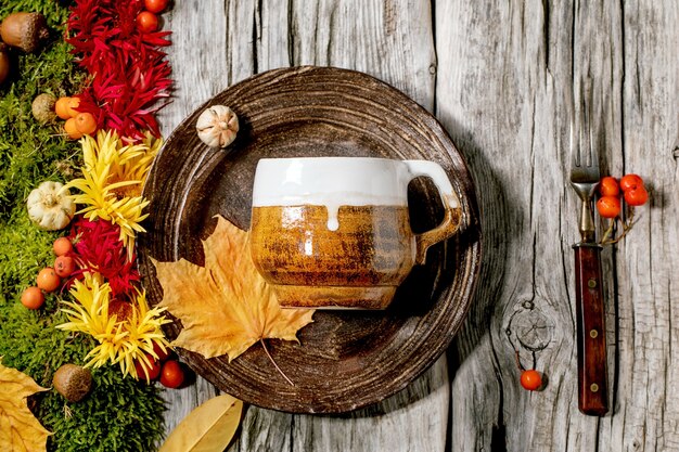 Autumn holidays table setting. Empty craft ceramic plate and mug on old wooden table decorated by fall yellow leaves, autumn berries, moss and flowers. Flat lay, copy space