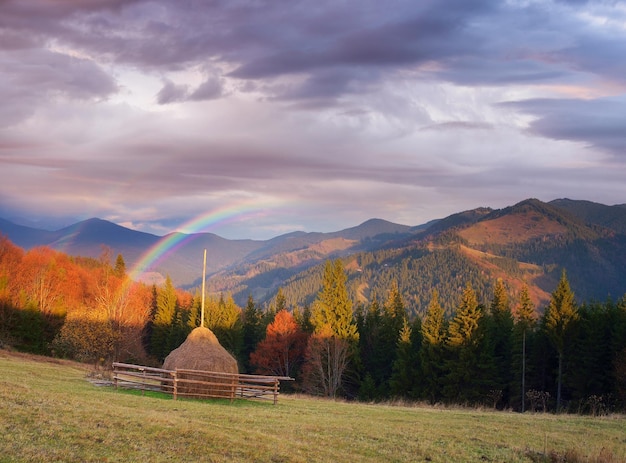 Autumn landscape in a mountain village. Haystack on the meadow. Rainbow over the hills. Collage of two frames. Art processing photos