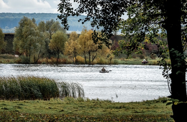 川と岸辺の木々を見下ろす秋の風景。池の真ん中でボートで釣り