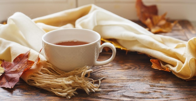 Autumn leaves with a cup of tea or coffee next to a scarf on a wooden background with copy space