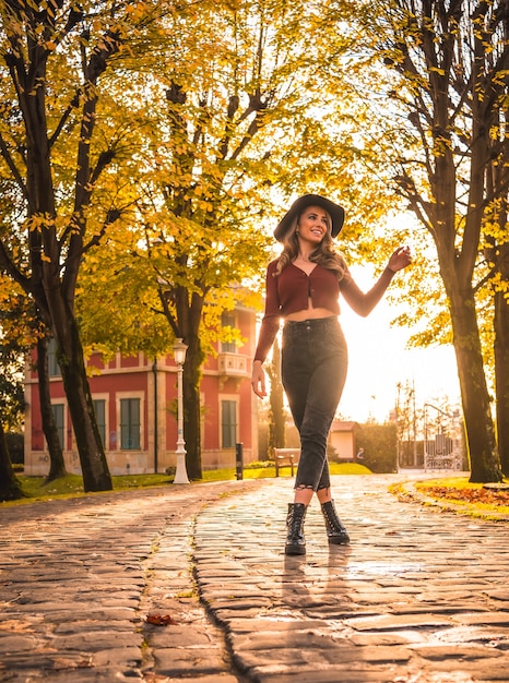 Autumn lifestyle at sunset blonde Caucasian woman in a red sweater and black hat enjoying nature in a park with trees Walking on the beautiful path of the city park