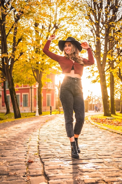 Autumn lifestyle at sunset blonde Caucasian woman in a red sweater and black hat enjoying nature in a park with trees Walking on the beautiful path of the city park