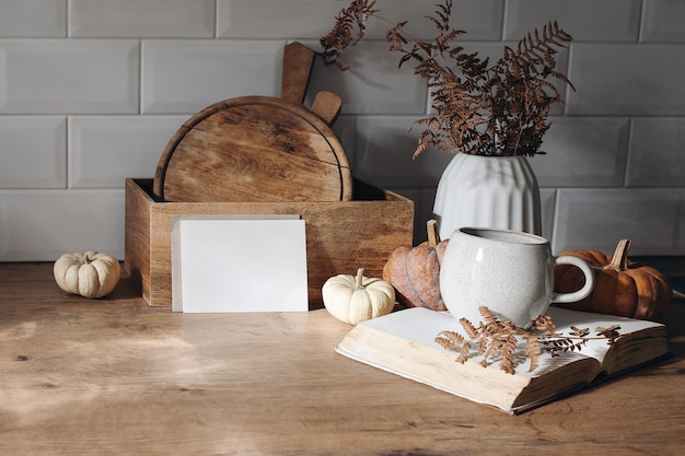 Autumn mockup scene Blank card open book cup of coffee wooden chop boards and dry fern leaves in sunlight Fall kitchen still life with pumpkins Wooden table background Thanksgiving Halloween