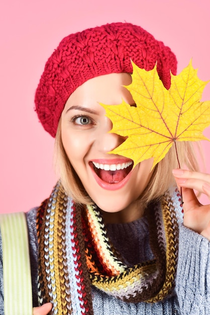 Autumn mood. Autumn time. Woman with autumn yellow maple leaf. Autumn fashion. Close up portrait of smiling woman with autumnal leaves. Woman hides eye by yellow leaf.