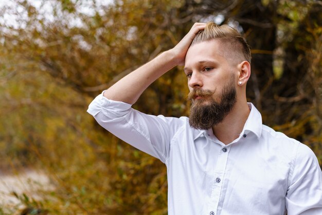 Autumn mood pensive young man in white shirt meditates and walks in park against the backdrop of bea...