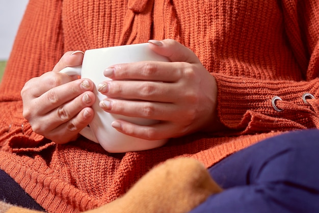 寒い秋の朝にさわやかなコーヒーを飲むオレンジ色のウールのプルオーバーの秋の気分の若い女性...