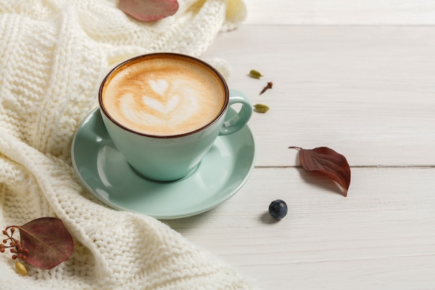 Autumn morning composition. Blue coffee cup with hot foamy cappuccino, cloves, cinnamon and warm sweater at white wood. Fall spicy drinks concept