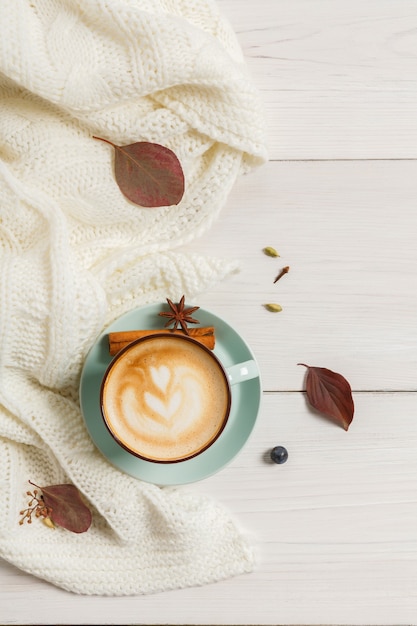 Autumn morning composition. Blue coffee cup with hot foamy cappuccino top view, cloves, cinnamon and warm sweater at white wood. Fall spicy drinks concept