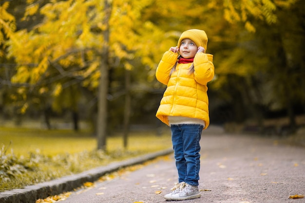 公園の秋。黄色い帽子と暖かい服を着たかわいい夢のような少女は、黄金の木々を背景に公園の路地に立って見上げています。秋の公園を歩いている笑顔の子供