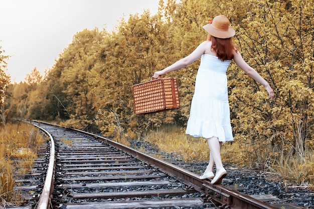 Фото Осенний парк девушка в белом сарафане и плетеном чемодане идет по рельсам