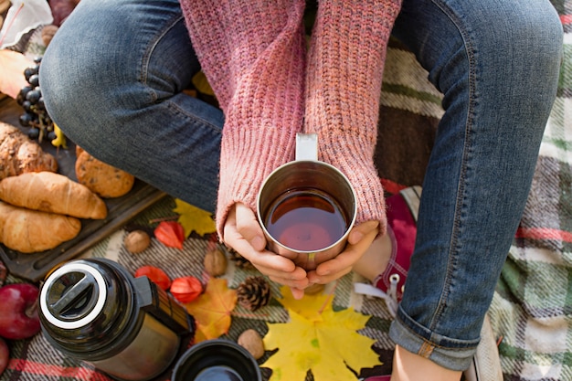 公園で秋のピクニック、暖かい秋の日。女の子は手にお茶の入ったカップを持っています。黄色の紅葉の毛布の上に花のバスケット。秋のコンセプト。