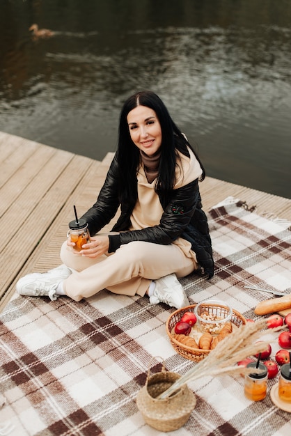 Autumn picnic on the shore stylish brunette girl sitting on a blanket and looking at the camera