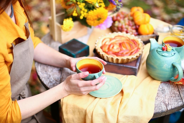 Picnic autunnale donna in abito giallo e grembiule di lino beve il tè dalla tazza al tavolo di legno in giardino bella tovaglia bollitore miele con cucchiaio torta di mele raccolta cachi uva foglia d'acero