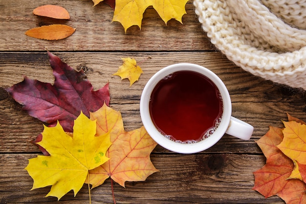 Maschera di autunno delle foglie gialle, una tazza di tè, una sciarpa e un pezzo di carta con la penna su fondo di legno