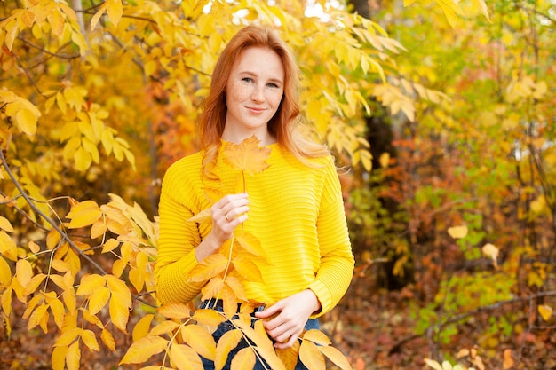 Осень. Портрет красивой счастливой рыжеволосой девушки в желтом джемпере среди осенних листьев. Цвет. Атмосфера. Место для текста. Фото высокого качества