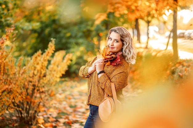 黄色の葉の花束を手にしたプラスサイズの女の子の秋のポートレート 秋の公園でスタイリッシュな女性