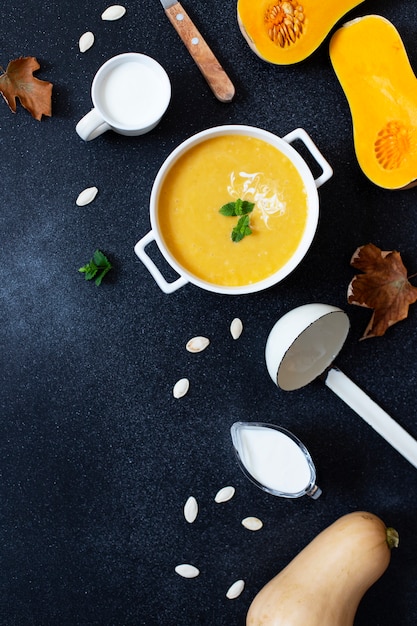 Autumn Pumpkin Cream soup on a black background, served with cream, pumpkin seeds and mint. Cut pumpkin, autumn leaves, soup ladle, white plate. Top view. Copyspace. Vertical