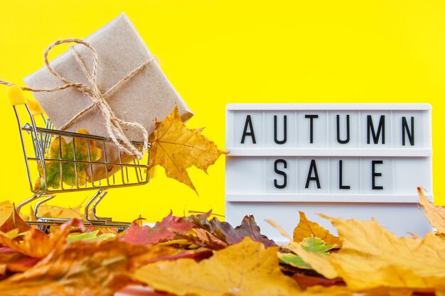 Foto autumn sale en discount concept winkelkarretje en gevallen gekleurde bladeren op gele achtergrond creatieve compositie voor reclame