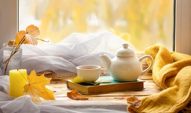 Фото Осенний натюрморт. чай дома. чайник, чашка и книга на фоне окна.