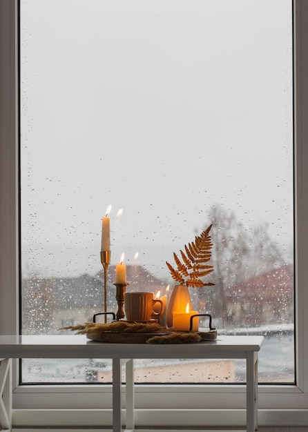Autumn still life with burning candles and a cup of coffee on the background of a window with raindr