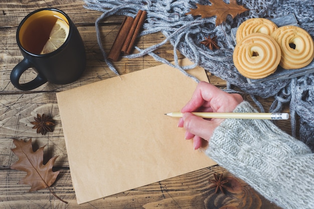 Autumn still life with cup of tea, cookies, sweater and leaves on wooden surface. concept of cozy autumn, Form for text. Female hand holding pen