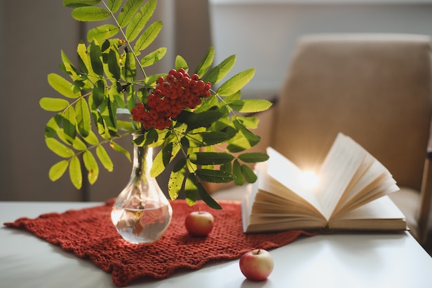 Autumn still life with a rowan branch in a vase and a book and apples in a cozy home interior