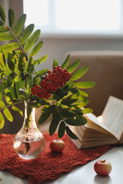 Autumn still life with a rowan branch in a vase and a book and apples in a cozy home interior