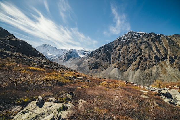 Autumn sunny landscape with highland valley and snow-covered high mountains under blue cloudy sky with cirrus clouds. atmospheric mountain scenery with sunlit valley among rocky mountains in autumn