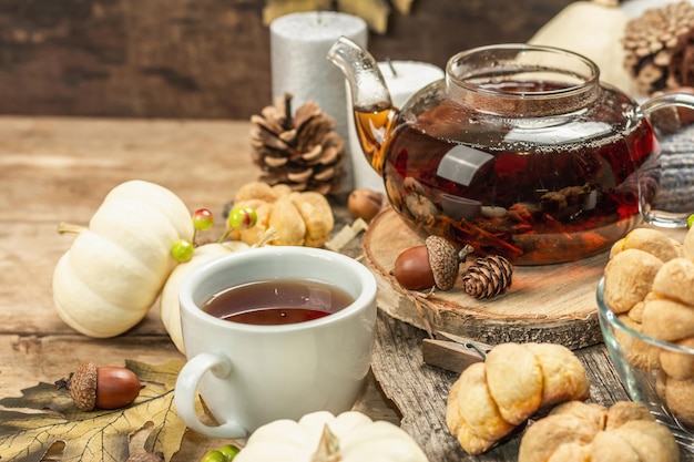 Autumn tea concept. Cookies with pumpkin puree, black tea in a glass teapot, fall decor. Wreath, candles, a cozy sweater. Old wooden background, copy space