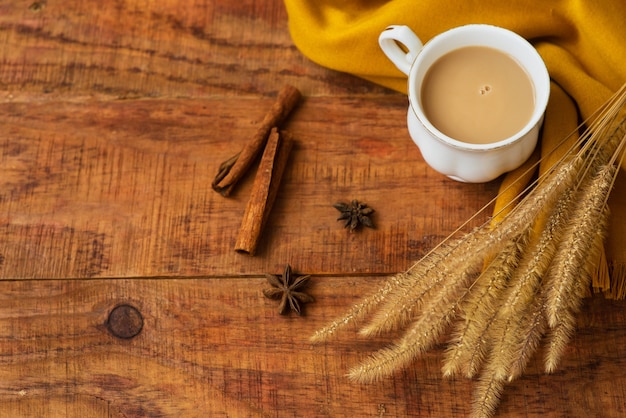 Осенняя композиция чашки чая с молоком, желтыми шарфами, палочками корицы и колосками пшеницы на деревянном фоне