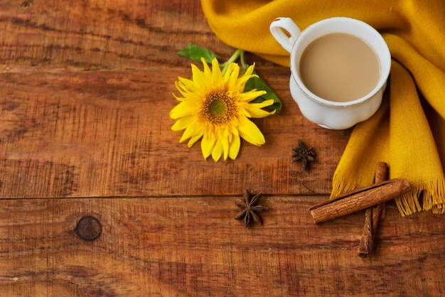 Autumn tea cup composition with milk, yellow scarfs, cinnamon sticks and sunflower on a wooden background. Autumn background. Warm, cozy atmosphere of autumn. Flat lay, layout. Place for text, frame