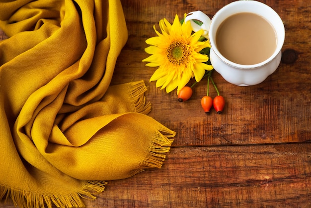 Composizione nella tazza da tè autunnale, sciarpa gialla, bacche di rosa e fiore di girasole su uno sfondo di legno