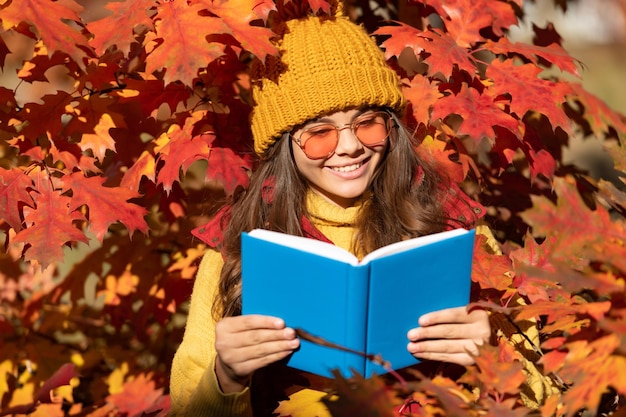 秋の10代の少女の肖像画秋の紅葉笑顔の子供は紅葉の背景に本を保持します