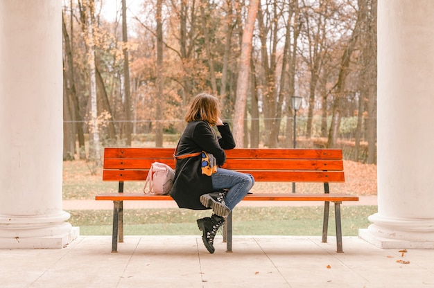 Autumn time. beautiful woman traveler dressed up in fall fashion. the girl sits on a bench and looks at the autumn landscape in the park. local solo travel concept