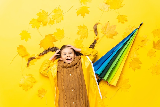 Photo autumn time for a little girl with pigtails the child is lying on yellow maple leaves with a multicolored umbrella in a raincoat and sweater