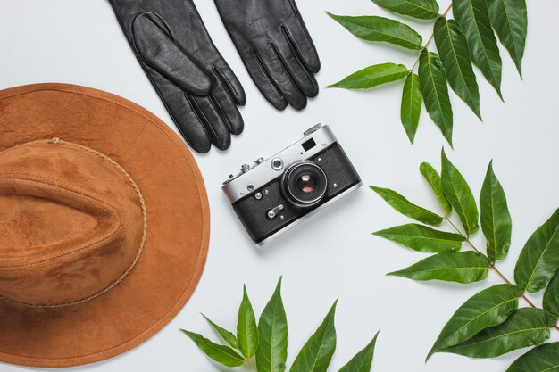 Autumn travel. Woman's accessories, retro camera on white background with green leaves. Felt hat, leather gloves. Top view