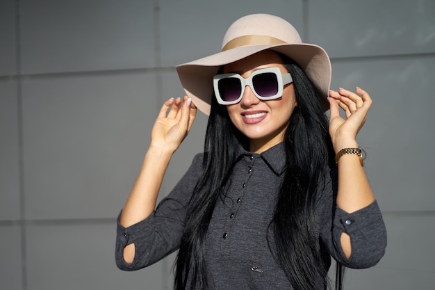 Autumn trendy collection. Happy beautiful brunette girl wearing stylish grey dress holding fashionable hat in the hands. Model in trendy outfit posing on grey wall. Outdoor portrait.