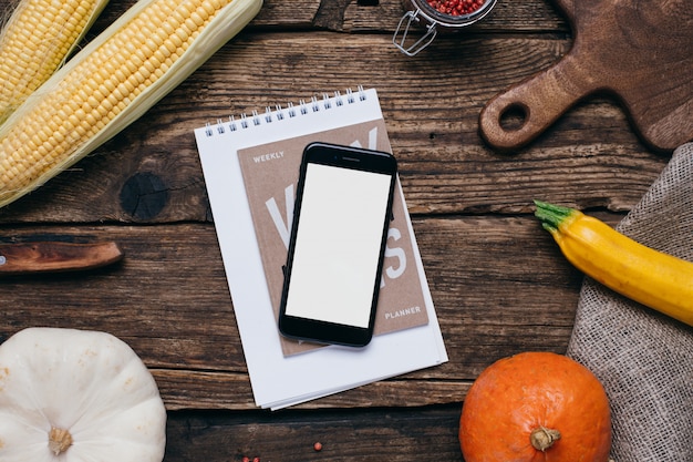 Foto verdure autunnali: cellulare con schermo bianco vuoto, zucche e mais con foglie gialle su legno