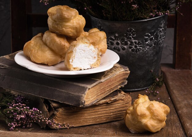 Autumn Vintage Provence Still Life with Group of French Dessert White Cream Eclairs on the Plate, Heather Flowers and Old Books. Gray Cement Rustic Rural Scenic Background near Rough Chair.