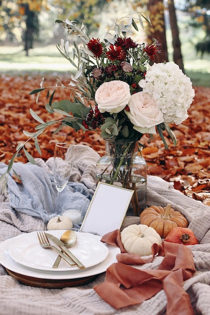 Autumn wedding table setting Garden party celebration picnic with golden cutlery porcelain platewine glass and white pumkins Rose flowers bouquet with olive branches Red beech leaves ground