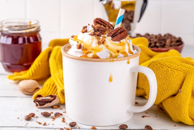 Autumn winter hot spicy drink Maple Pecan coffee latte with whipped cream cup on white wooden table with cozy sweater and ingredients copy space