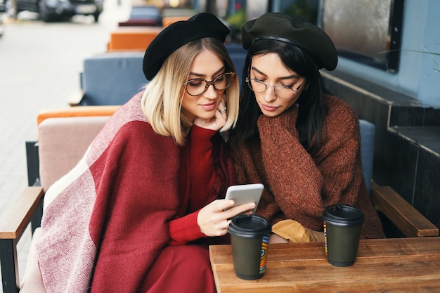 Autumn winter portrait of two young women bloggers in an outdoor cafe looking to smartphone screen, drinking coffee to go, talking.