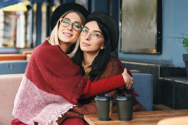 Autumn winter portrait of two young women in an outdoor cafe, drinking coffee to go, talking.