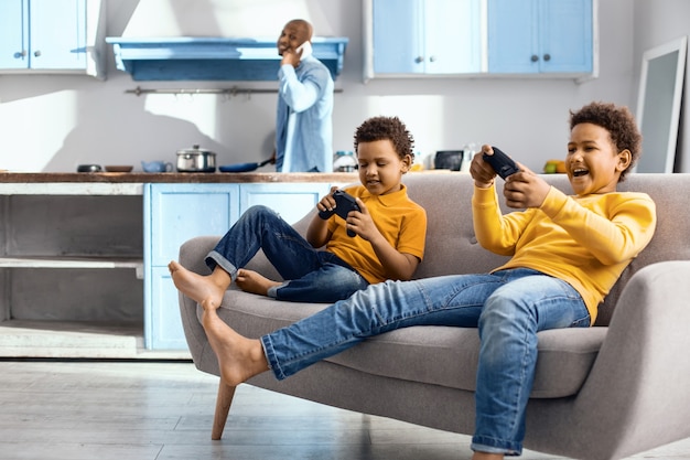 Avid gamers. Cheerful little boys sitting on the sofa and being immersed into playing a video game with controllers while their father cooking for them in the background