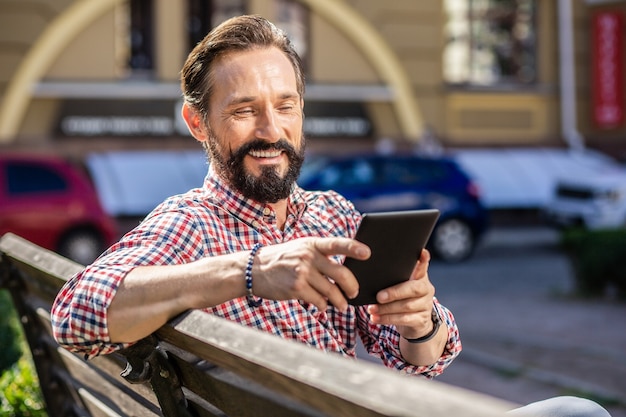 Avid reader. Cheerful bearded man sitting on the bench while reading an e-book