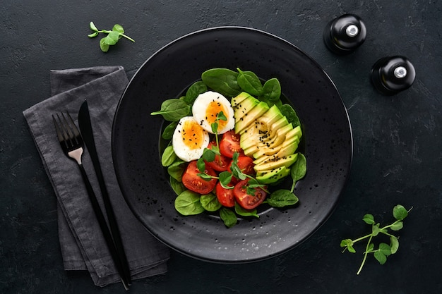 Avocado, cherry tomato, spinach and chicken egg, microgreens peas and black sesame seeds fresh salad in black bowl on black slate, stone or concrete background. Healthy food concept. Top view.