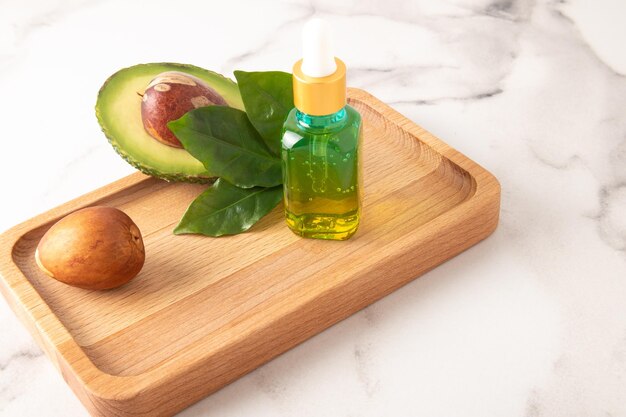 Avocado extract in a colored bottle on a wooden tray with ripe avocado moisturizing vitamin E marble background