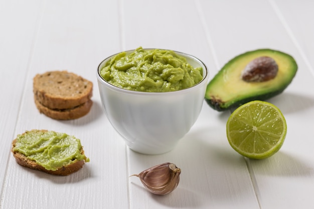 Avocado, lime, bread and garlic with a bowl of guacamole on a white table. Diet vegetarian Mexican food avocado. Raw food.