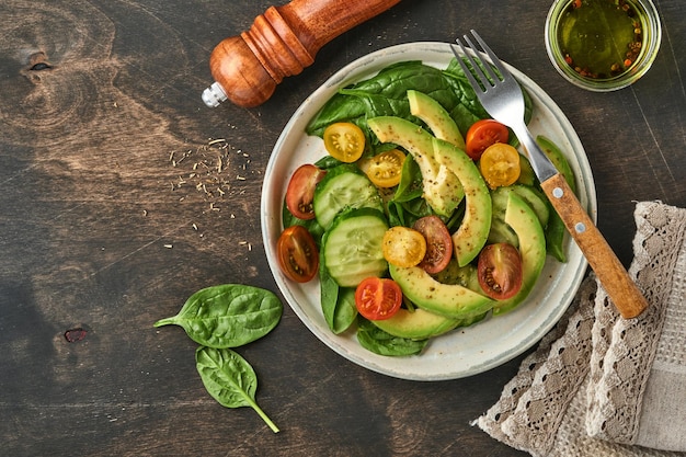 Avocado, red, yellow, black cherry tomato, spinach and cucumber fresh salad with spices pepper and olive oil in grey bowl old wooden table background. Healthy food concept. Top view.